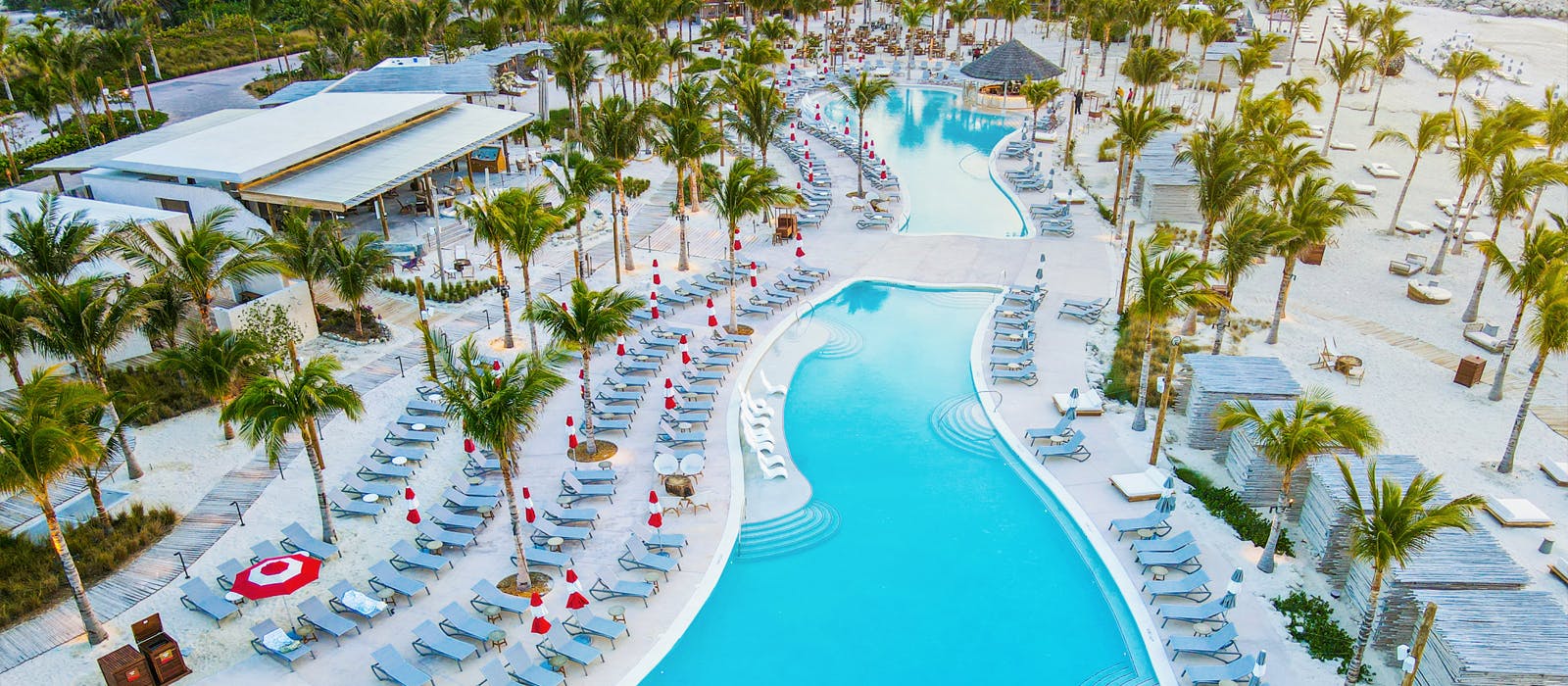 Two lagoon-shaped pools surrounded by palm trees and lounge chairs, next to a beach.